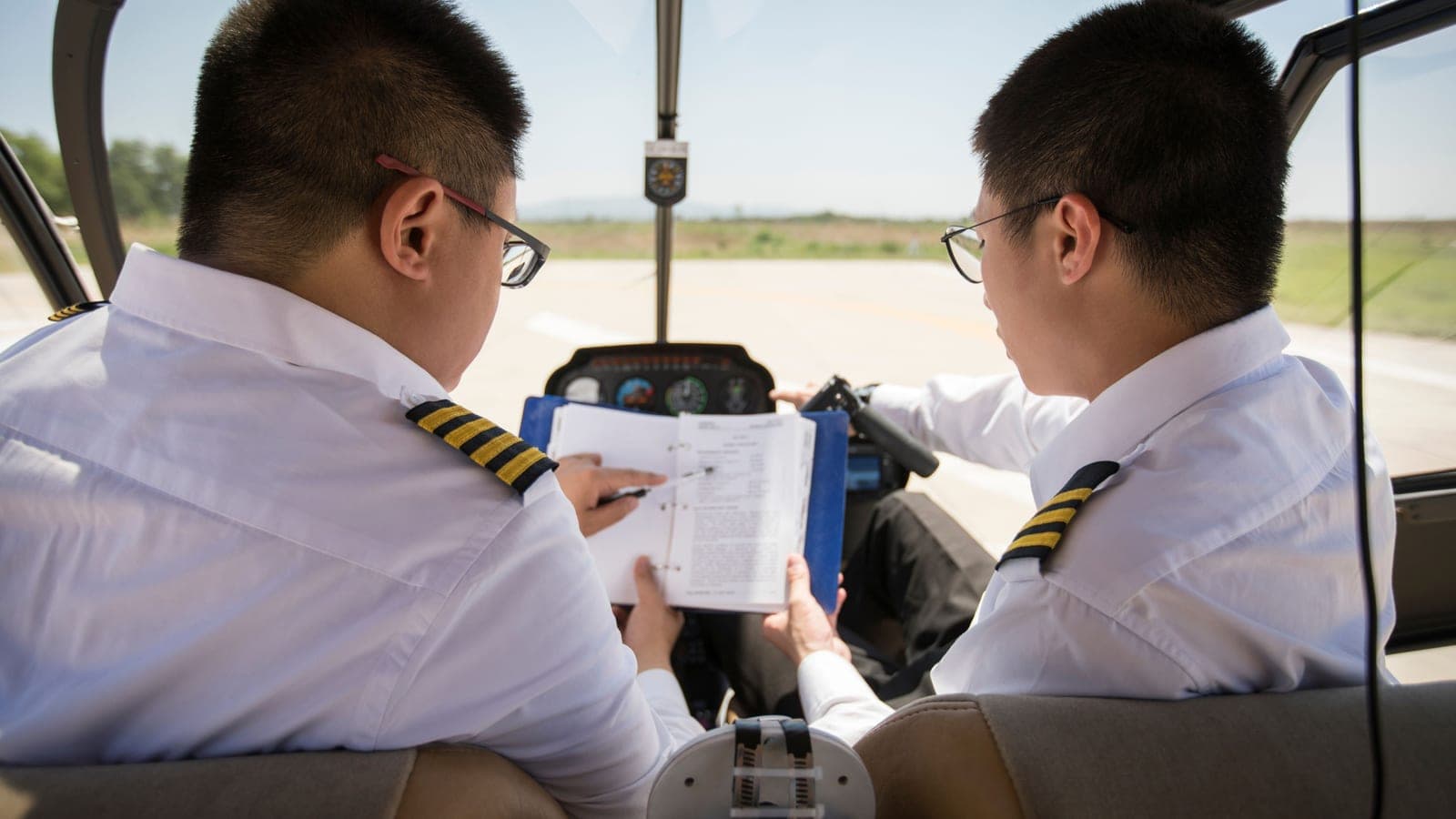 Two pilots seen from behind in a light aircraft cockpit, one holding an open ring-bound checklist across the yoke