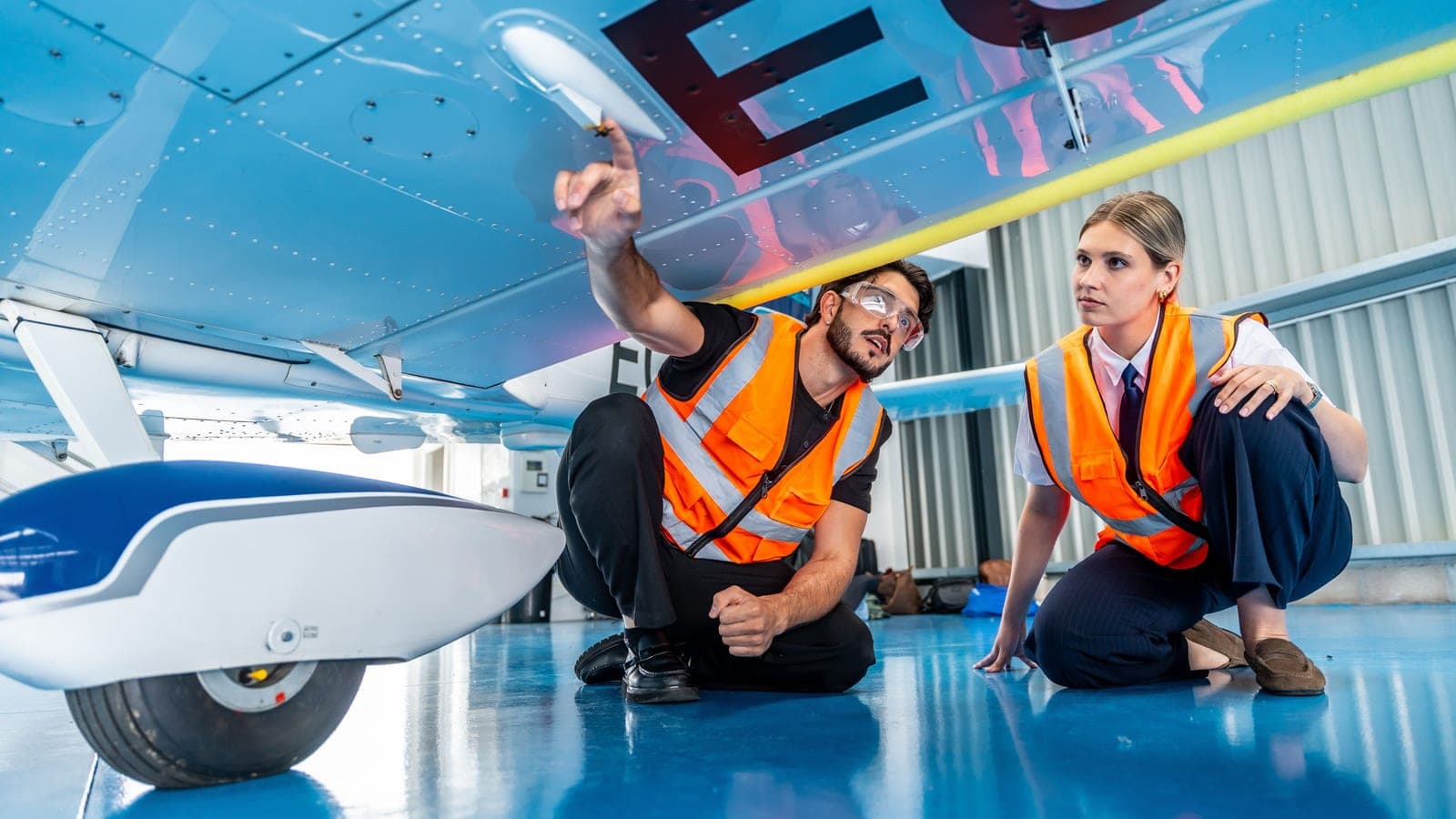Two engineers in hi-vis vests crouched beneath the wing of a light aircraft in a hangar, one pointing up at the underside