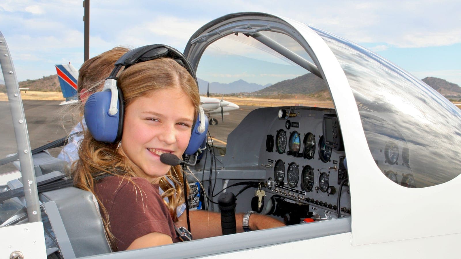 A young student in ear defenders and a headset grinning from the open cockpit of a light aircraft