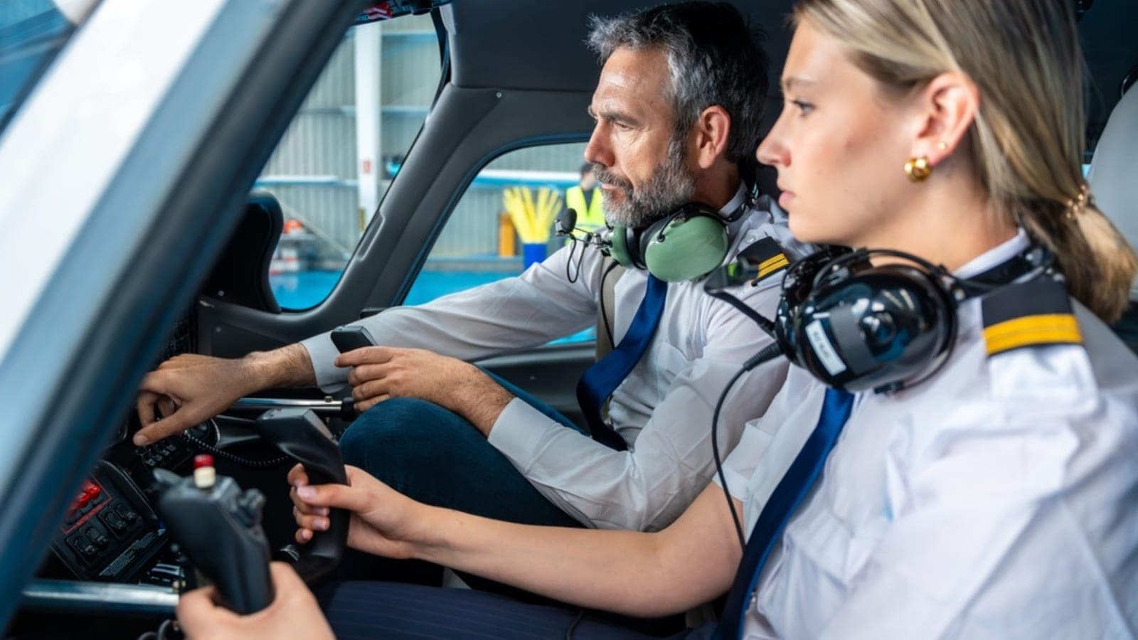 An instructor and a student side by side in a light aircraft cockpit, both wearing headsets, hangar visible through the windscreen
