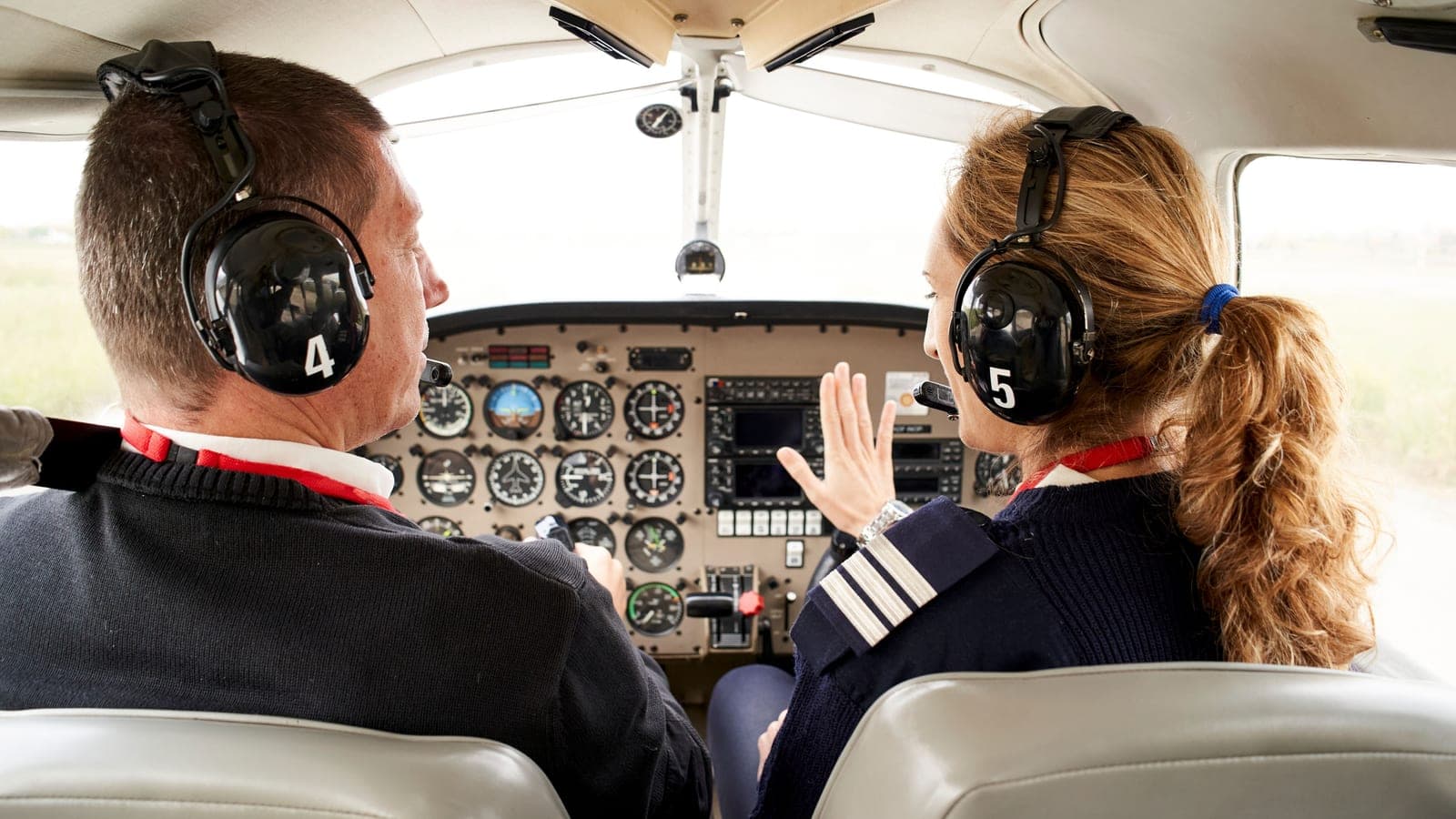 Two pilots from behind at a full analog instrument panel, one gesturing toward the gauges as the other flies