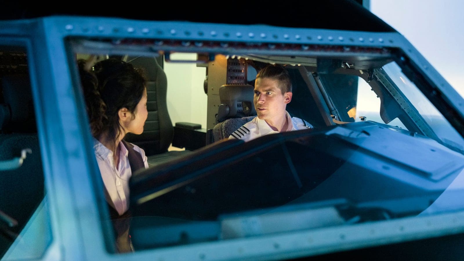 Two pilots in conversation in a dimly lit cockpit, seen through the windscreen in cool blue light