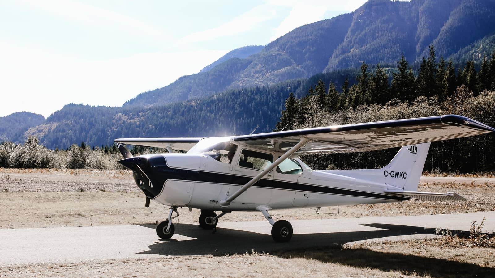 A Cessna 172 parked on a remote ramp with forested mountains rising behind it