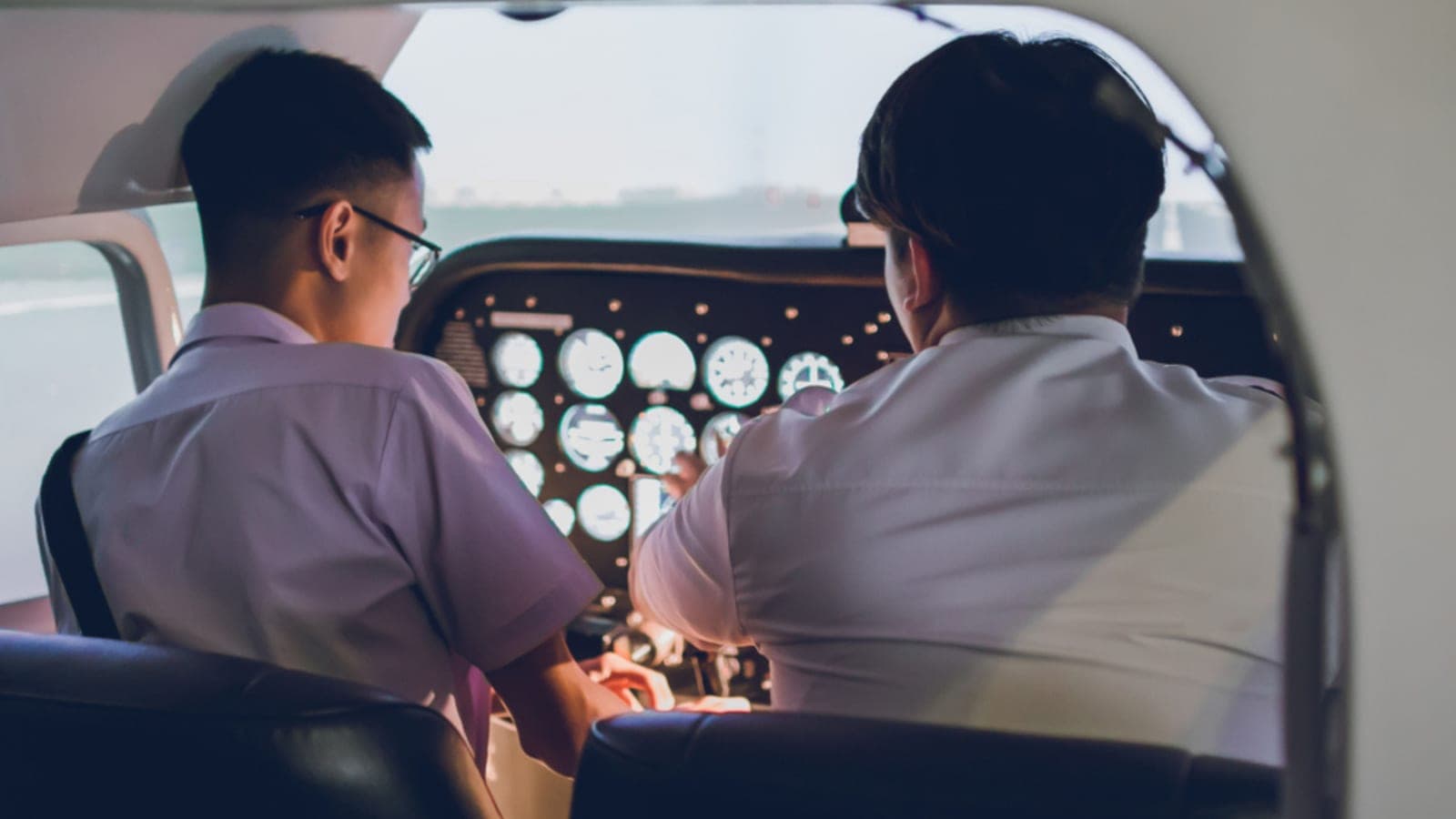 Two student pilots seated at a vintage analog instrument panel, seen from behind during a lesson
