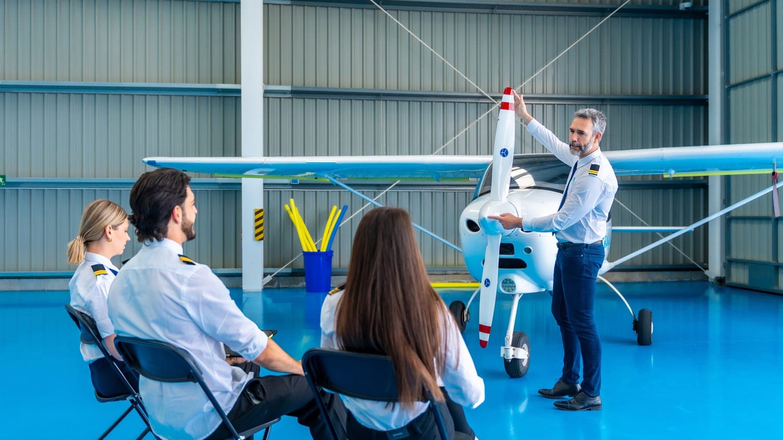 Three students seated in a bright hangar watching an instructor present a light aircraft